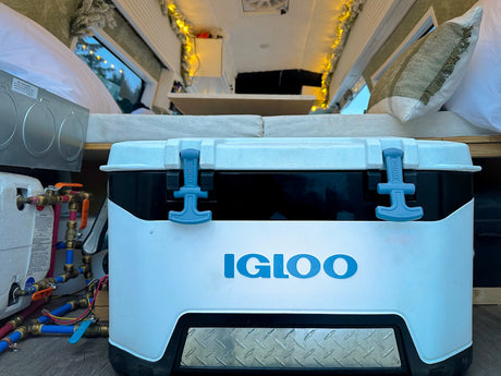 Igloo cooler and utilities within a cozy RV rental. Van life essentials, from plumbing to comfy bed, glow with string lights.