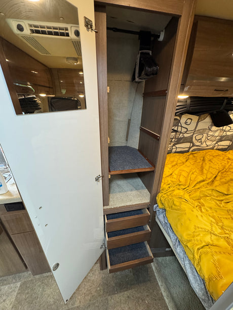 RV rental storage: Open closet with hanging rod, shelves, and carpet-lined drawers. A bed with a yellow blanket is visible nearby.