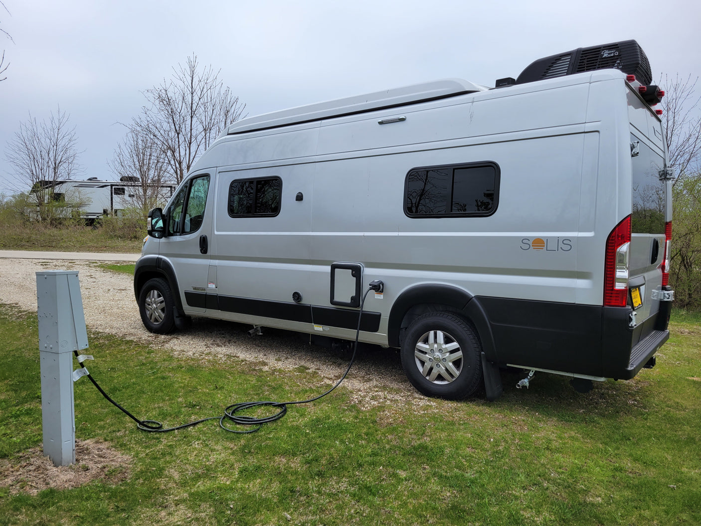 RV rental Winnebago Solis camper van (silver) plugged into a power pedestal at a grassy campsite, ready for camping.