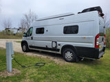 RV rental Winnebago Solis camper van (silver) plugged into a power pedestal at a grassy campsite, ready for camping.