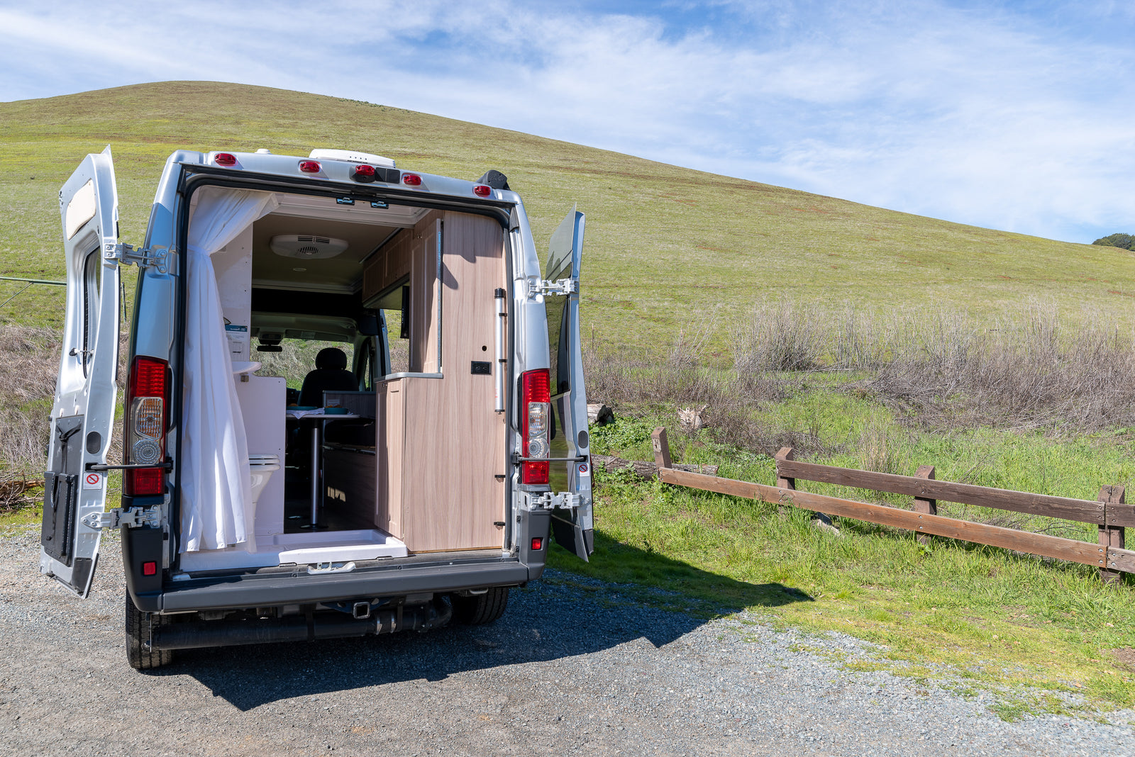 RV rental camper van with open rear doors, revealing its functional interior with a toilet, kitchen, and person inside. Parked by a green hill.