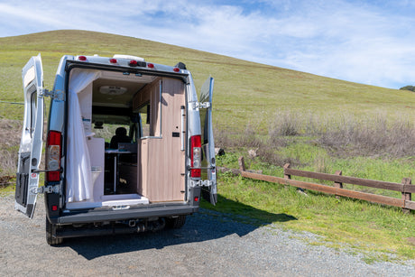 RV rental camper van with open rear doors, revealing its functional interior with a toilet, kitchen, and person inside. Parked by a green hill.