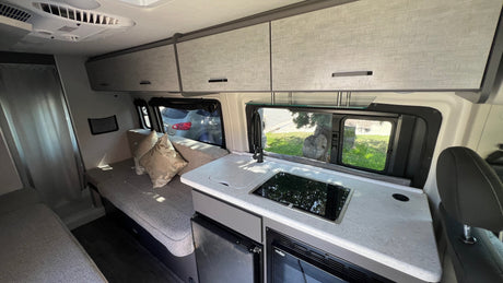 Inside an RV rental: Compact kitchen with sink & cooktop, overhead storage, and lounge seating. Ideal for travel.