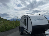 Bushwhacker teardrop camper RV rental parked with snowy mountains and cloudy skies in background.