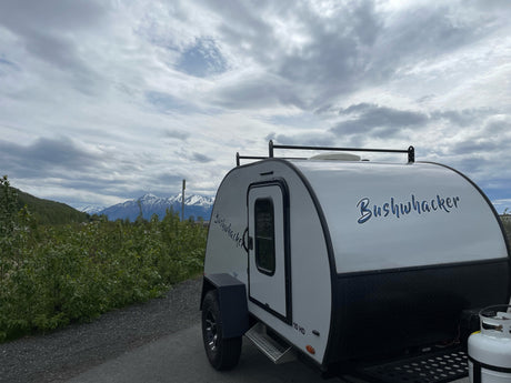 Bushwhacker teardrop camper RV rental parked with snowy mountains and cloudy skies in background.