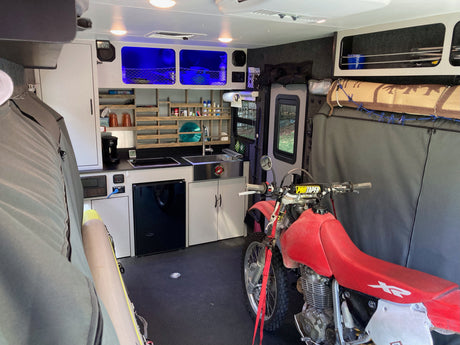 Interior of an rv rental toy hauler, featuring a dirt bike secured alongside a compact kitchen, storage, and ambient blue lighting.