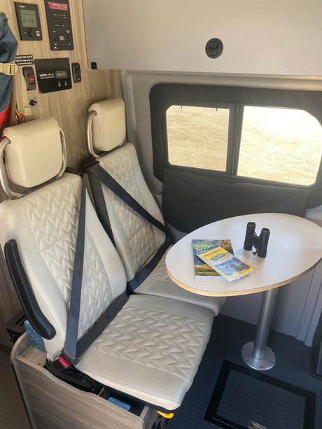 Comfortable RV rental interior featuring passenger seating, a dining table with guides, and control panels for travel.
