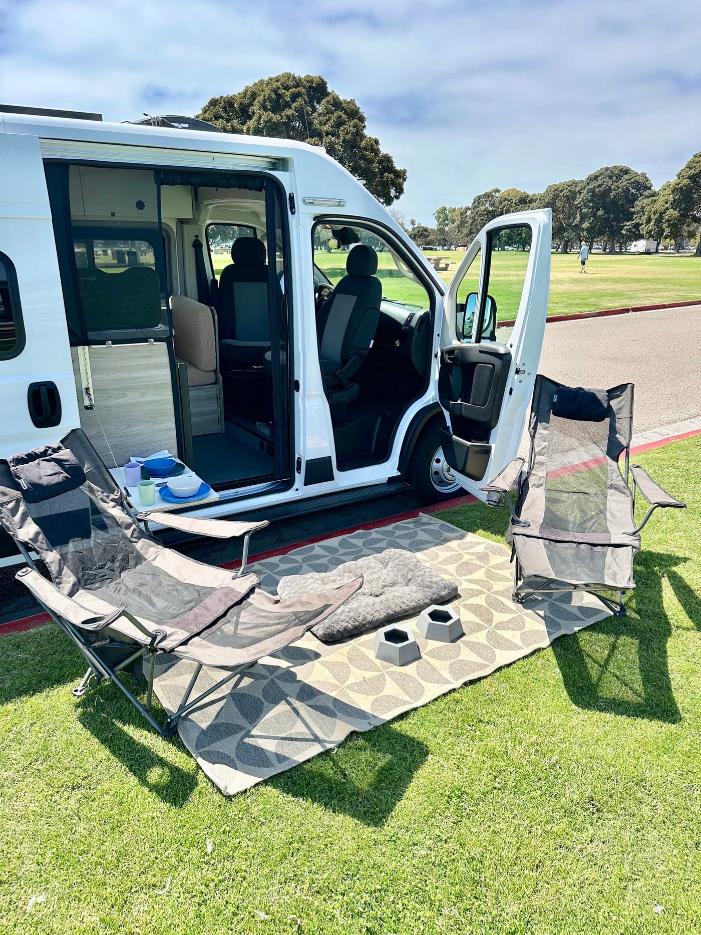 RV rental experience: White camper van open to a cozy outdoor setup on a rug with chairs, pet bed, and bowls in a park. — photo 12