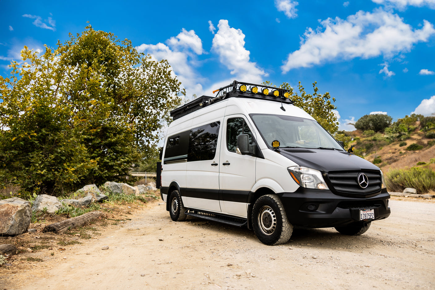 Off-road equipped white Mercedes Sprinter RV rental van parked on a dirt path, ready for adventure under a blue sky.