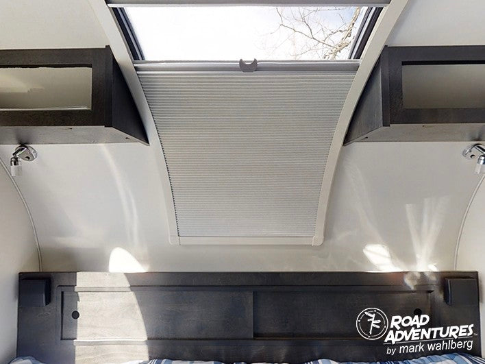 Inside an RV rental: a bright skylight with a pleated shade, dark wood overhead cabinets, and reading lights above a headboard.