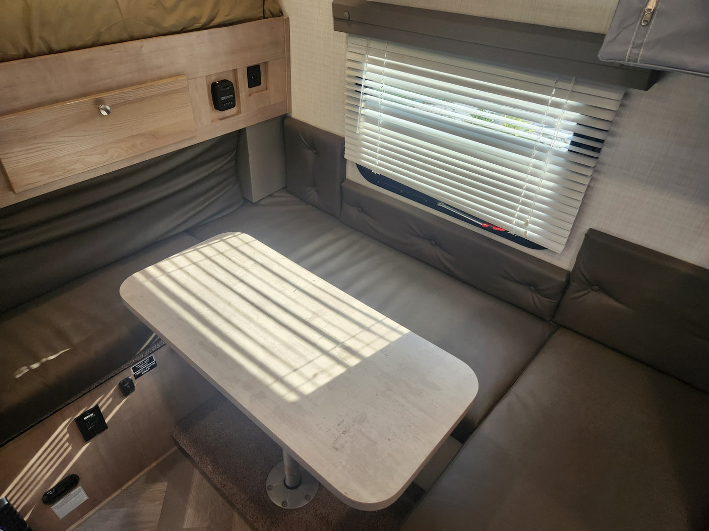 Bright RV rental interior: a comfortable dinette with light wood table, dark benches, and sunlit window blinds.