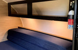 Inside an RV rental: blue bed, overhead cabinets, under-cabinet lighting, and a fire extinguisher for safety.