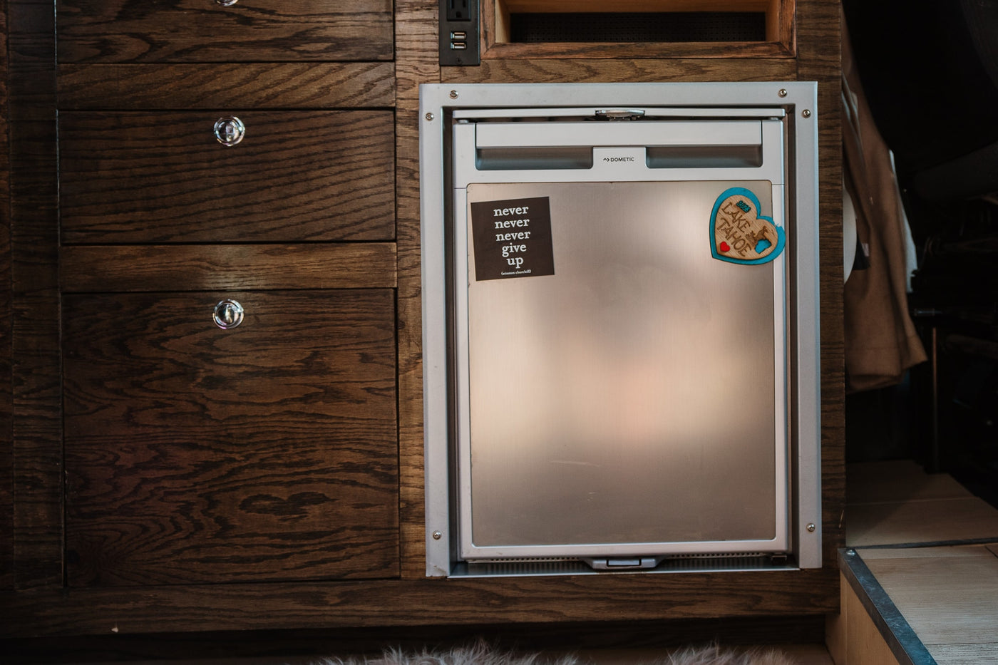 An RV rental's Dometic compact fridge & dark wood cabinets. Features a "Never Give Up" sticker and Lake Tahoe magnet. — photo 6
