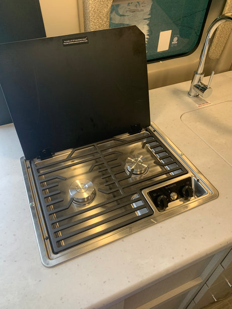 RV rental kitchen with a Dometic 2-burner gas cooktop, its black cover open, showing grates and controls, next to a sink.