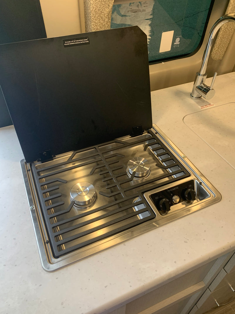 RV rental kitchen with a Dometic 2-burner gas cooktop, its black cover open, showing grates and controls, next to a sink. — photo 18