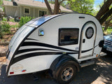 A white T@G teardrop camper, a potential RV rental, features black wavy stripes, a round door, and a solar panel.