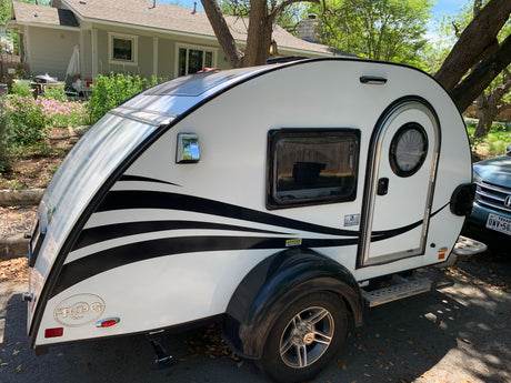 A white T@G teardrop camper, a potential RV rental, features black wavy stripes, a round door, and a solar panel.