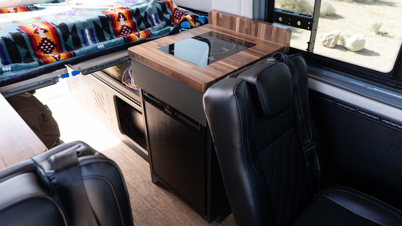 RV rental interior: cozy bed with patterned blanket, compact kitchen with induction cooktop & fridge, black seats. — photo 2