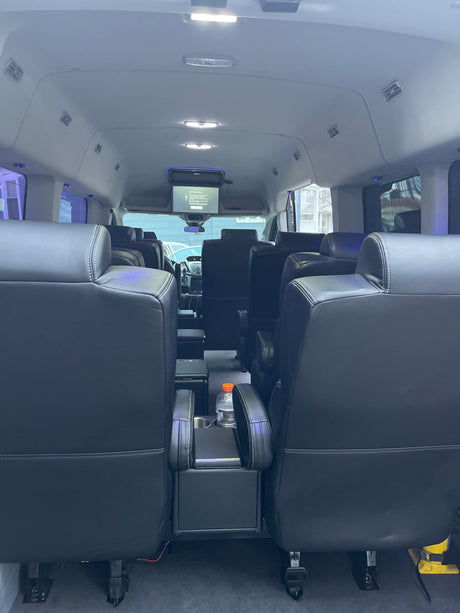 Comfortable interior of a passenger van, ideal for RV rental. Features plush black leather seats, overhead screen, and ample lighting.