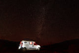 White RV rental camper van with glowing interior lights under a brilliant starry night sky with distant mountains.