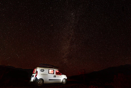 White RV rental camper van with glowing interior lights under a brilliant starry night sky with distant mountains.