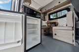 Inside an rv rental: an open refrigerator shows shelves, alongside a cozy dinette, wood cabinetry, and a window view.