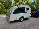 A white & green teardrop RV rental, perfect for outdoor travel, is hitched to an SUV on a gravel path surrounded by trees.