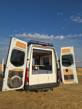 RV rental van, rear doors open revealing custom interior: bed, table, sink. Storage pockets on doors, roof rack above.