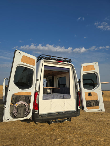 RV rental van, rear doors open revealing custom interior: bed, table, sink. Storage pockets on doors, roof rack above.