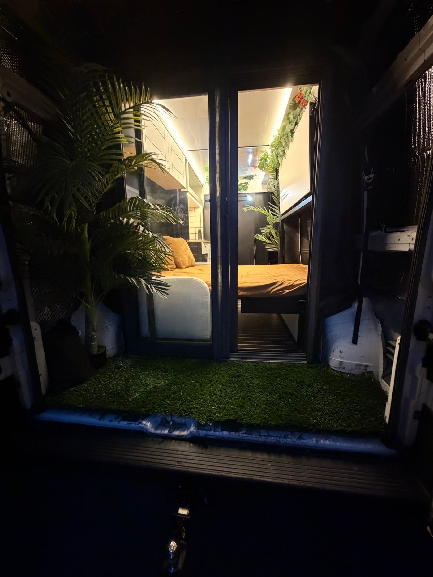Night view of an RV rental's inviting interior: two beds, plants, strip lighting, and a soft artificial grass entrance. — photo 5