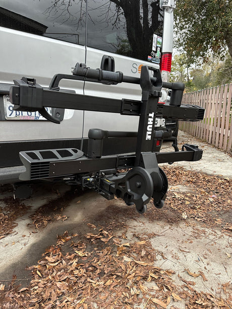 Rear view of a gray RV rental, featuring a folded Thule bike rack. Leaves cover the concrete ground.
