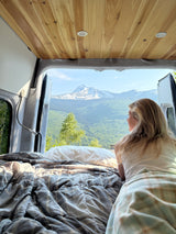 Stunning mountain views from a cozy rv rental bed. A blonde woman relaxes, enjoying the serene van life experience.
