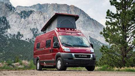 Red pop-top camper van parked on a dirt road with mountains & trees.