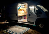 Nighttime view of a cozy campervan RV rental with its door open, revealing a lit interior, bed, and hammock.