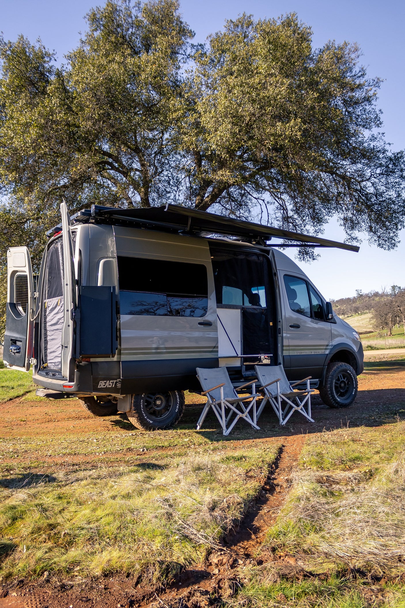 Overland camper van, ideal for an RV rental getaway, parked under trees with awning out & chairs set for relaxing.