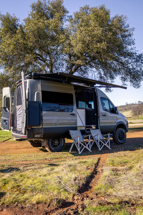 Overland camper van, ideal for an RV rental getaway, parked under trees with awning out & chairs set for relaxing.