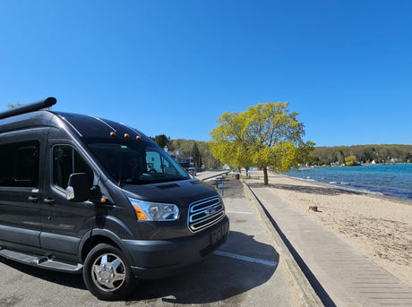 Enjoy the view from this RV rental, a dark Ford Transit van, parked by a sunny beach with blue water and green trees.