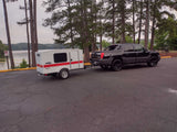 Experience nature with this RV rental: a black truck towing a compact white Runaway teardrop camper by a lake.