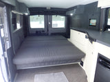 RV rental camper van interior showing a large grey bed, light walls, dark counters, and a wall-mounted screen.