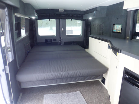 RV rental camper van interior showing a large grey bed, light walls, dark counters, and a wall-mounted screen.