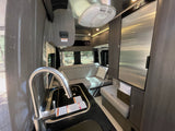 Spacious RV rental interior featuring a modern kitchen with sink & stove, sleek fridge, & comfortable lounge area with forest views.