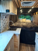 Cozy RV rental interior with a bed, compact kitchen with sink, wood paneling, string lights, and a window view.