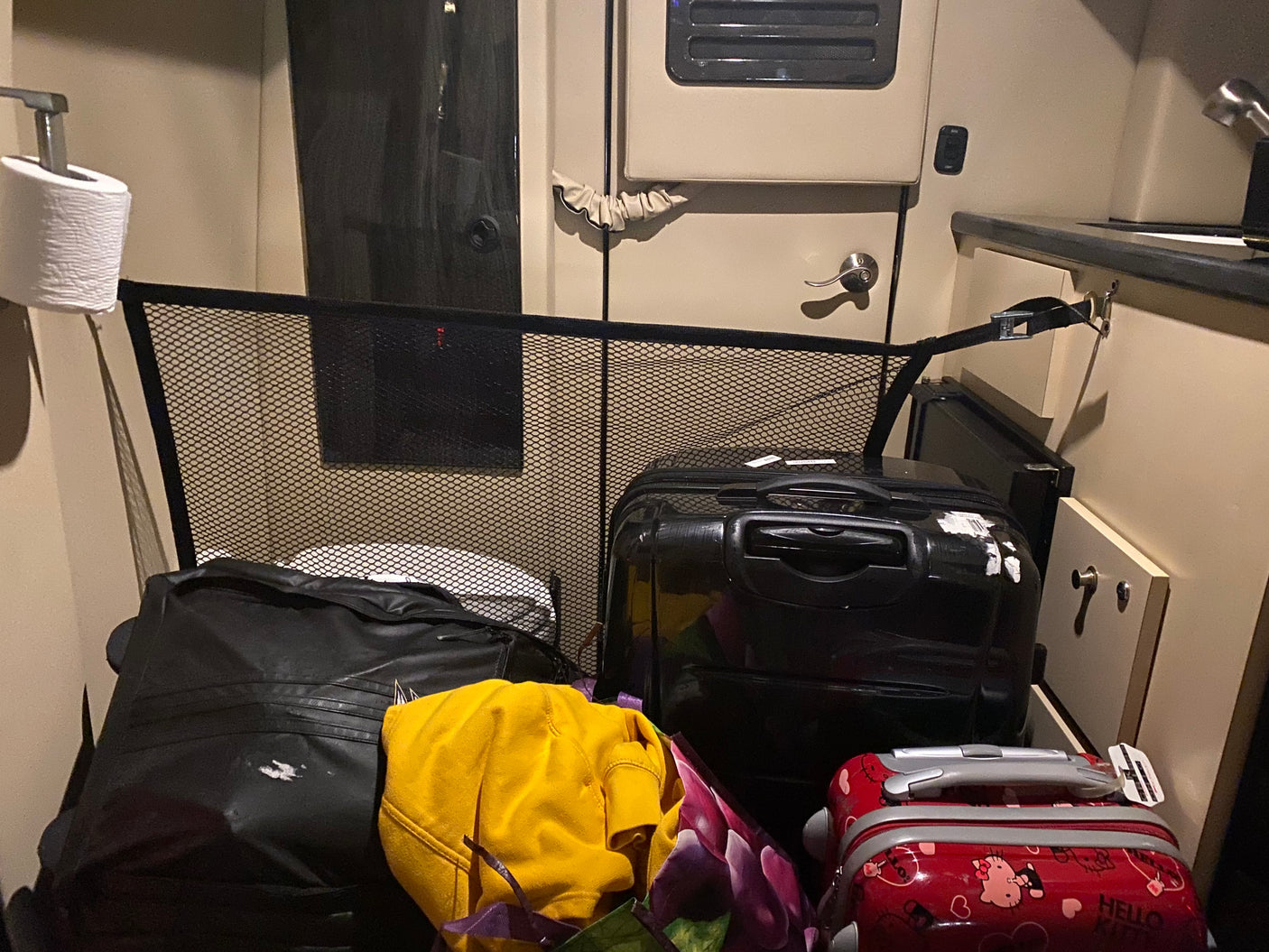 Inside an RV rental: a roll of toilet paper on the left, a black mesh barrier, and multiple suitcases with clothes filling the compact interior.