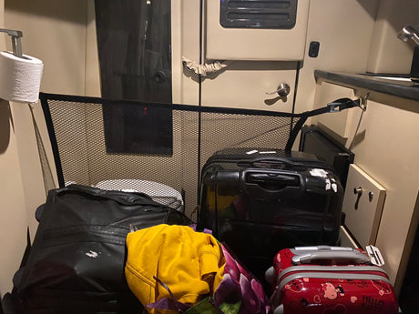 Inside an RV rental: a roll of toilet paper on the left, a black mesh barrier, and multiple suitcases with clothes filling the compact interior.