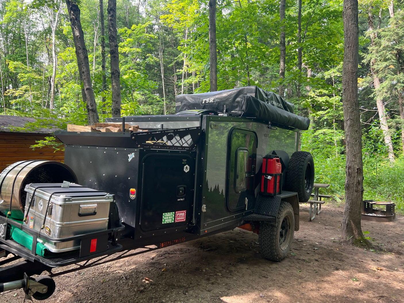 An overland trailer RV rental by Boreas Campers sits in a wooded campsite, ready for adventure with a rooftop tent.