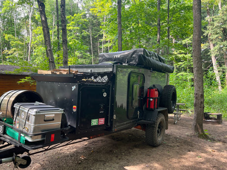 An overland trailer RV rental by Boreas Campers sits in a wooded campsite, ready for adventure with a rooftop tent.