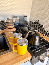 RV rental kitchen: pour-over coffee maker, gooseneck kettle, two yellow mugs, stove, and a stylish black/white tile backsplash.