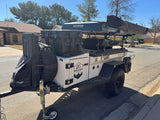 A sturdy white overland camper trailer with a dark blue rooftop tent and gear, parked on a street, perfect for an RV rental adventure.