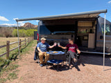 A couple toasts by their RV rental, enjoying snacks under an awning with scenic mountains in the background.