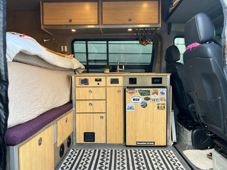 RV rental interior: cozy bed, custom bamboo kitchen with sink & fridge, plus front seats. Ideal for van life adventures!